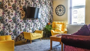 Un salon avec un papier peint à motifs floraux, un téléviseur à écran plat, un fauteuil jaune, une table basse en bois et un canapé violet.