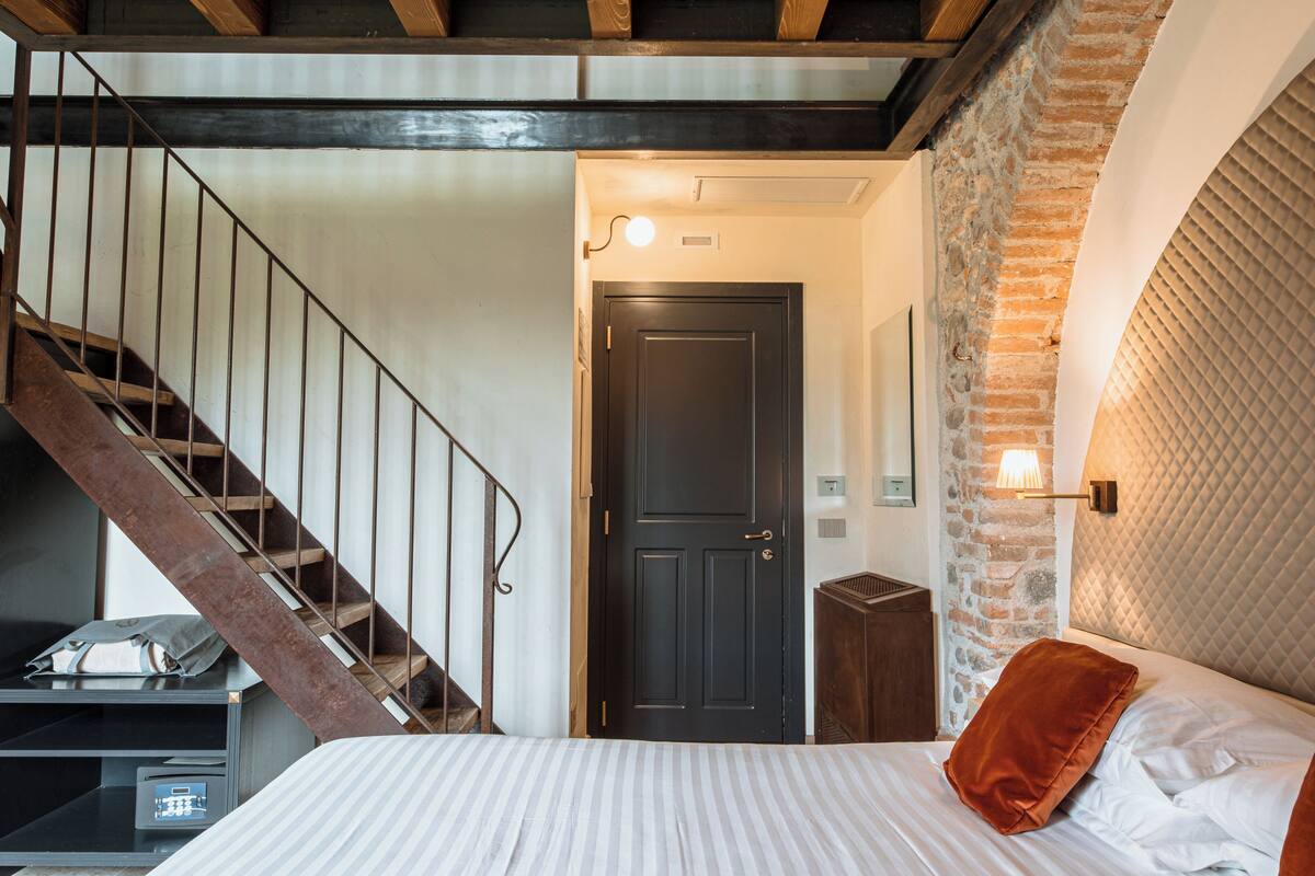 Une chambre à coucher avec un lit, un escalier en bois, une porte noire et un mur en pierre.