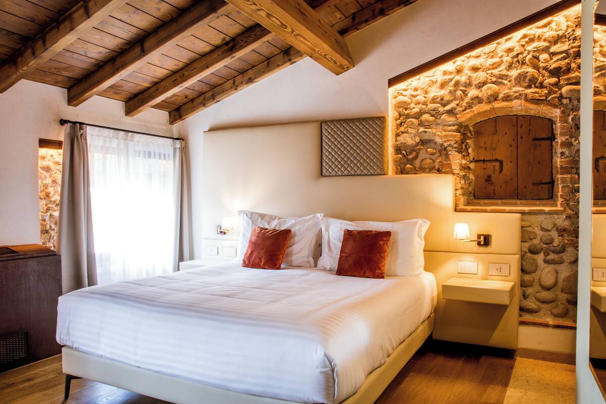 Une chambre avec un grand lit, des poutres en bois au plafond, un mur en pierre et une cheminée en pierre.