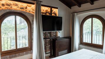 Un dormitorio con una cama grande, un televisor montado en una pared de piedra y dos ventanas amplias con cortinas.