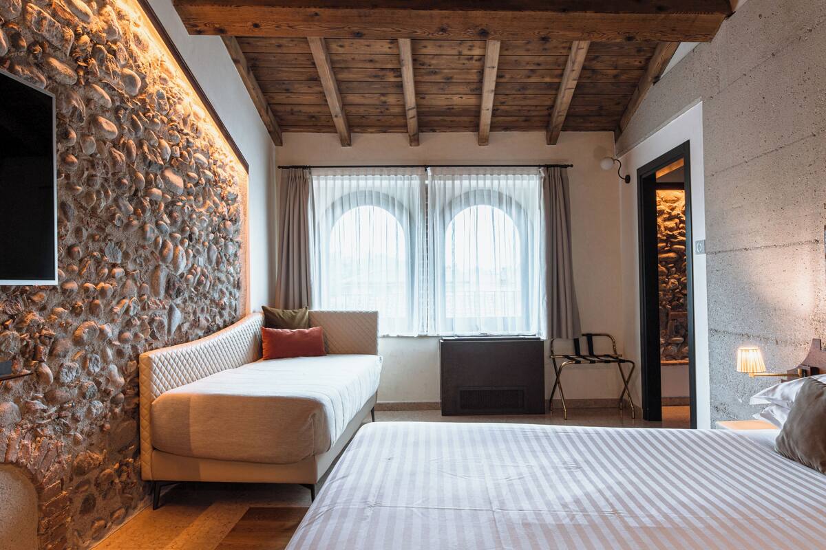 Une chambre avec un mur en pierre, des poutres en bois au plafond, un lit, un canapé, une cheminée et une fenêtre avec des rideaux.