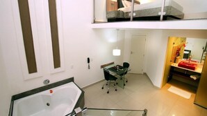 Une chambre d’hôtel moderne dotée d’un balcon fermé par des baies vitrées, d’une salle de bain séparée avec un lavabo rouge, et d’un coin repas avec une table et des chaises en verre.