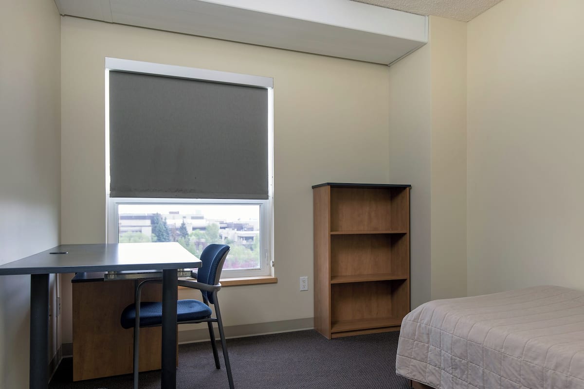 Une petite pièce, simplement aménagée, avec un bureau, une chaise, une bibliothèque et un lit.