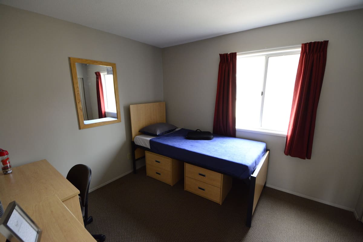 Une chambre avec un lit simple, un bureau, une chaise, un miroir et une fenêtre avec des rideaux.