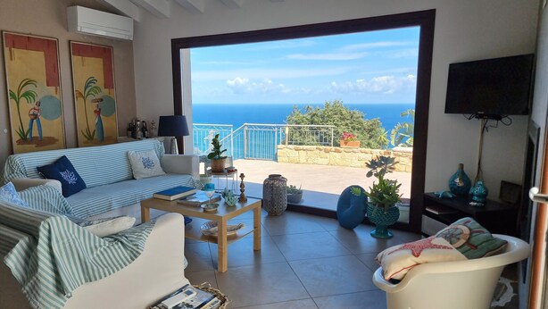 A living room with a sofa, coffee table, and chairs, a TV, and a view of the sea.