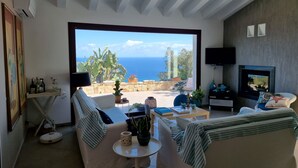 A modern living room with a large window overlooking the sea, a sofa, a coffee table, and a TV.
