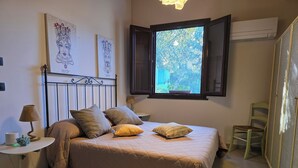A bedroom with a bed, a bedside table, a lamp, a chair, and a window with an open shutter.