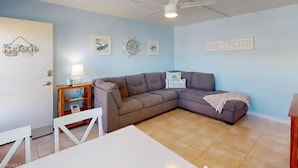 Un salon avec un grand canapé gris en forme de L, une table basse blanche, une petite table d’appoint en bois avec une lampe et un ventilateur de plafond.