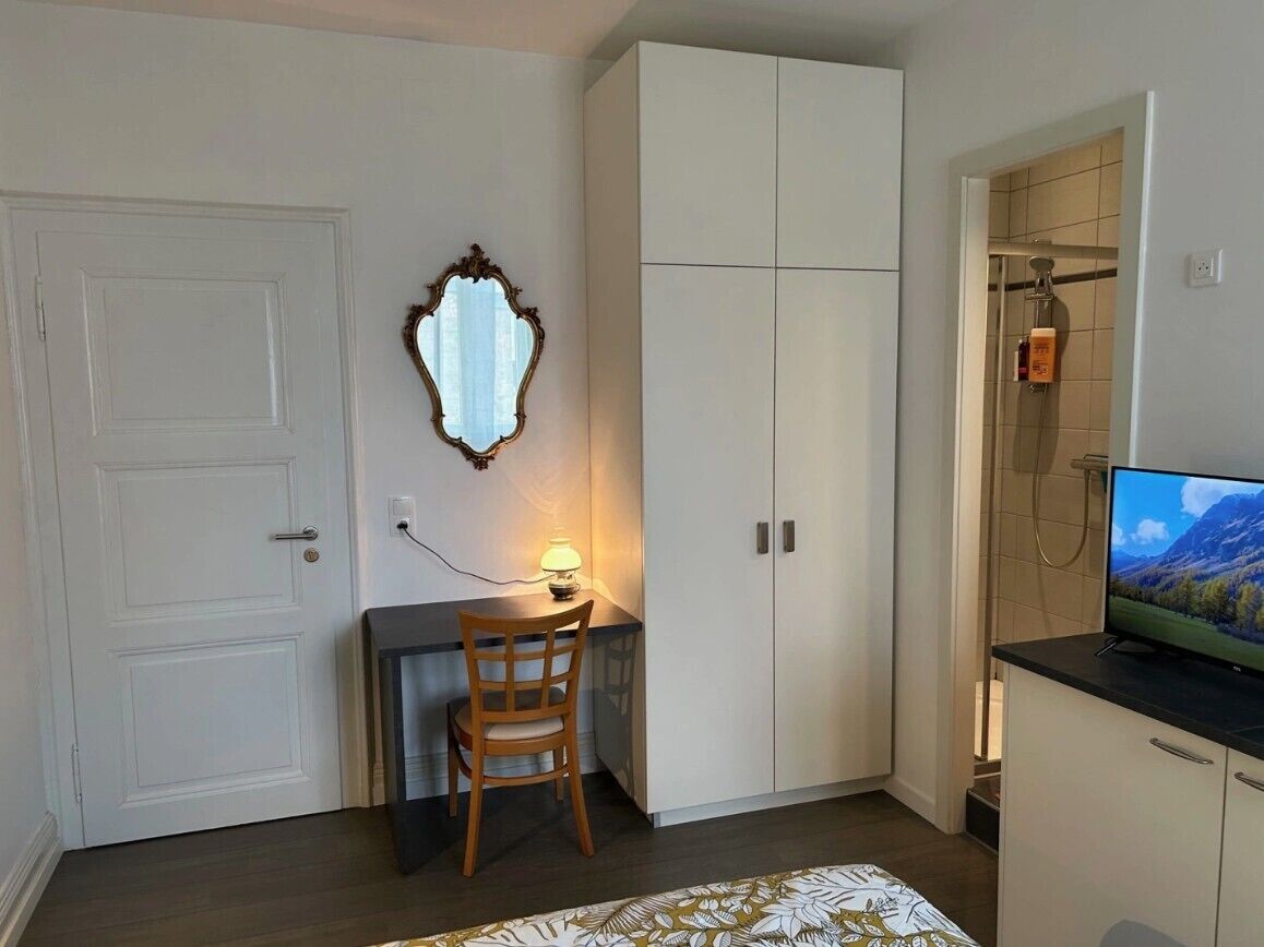 Ein kleines Zimmer mit Schreibtisch, Stuhl, Kleiderschrank und Fernseher.
