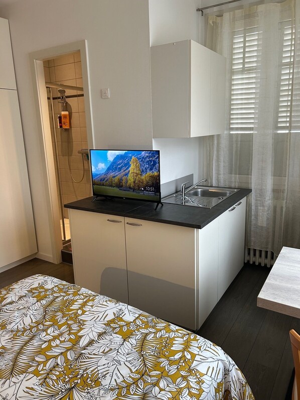 Habitación de hotel compacta con kitchenette, ducha, cama con colcha estampada y ventana con cortinas.