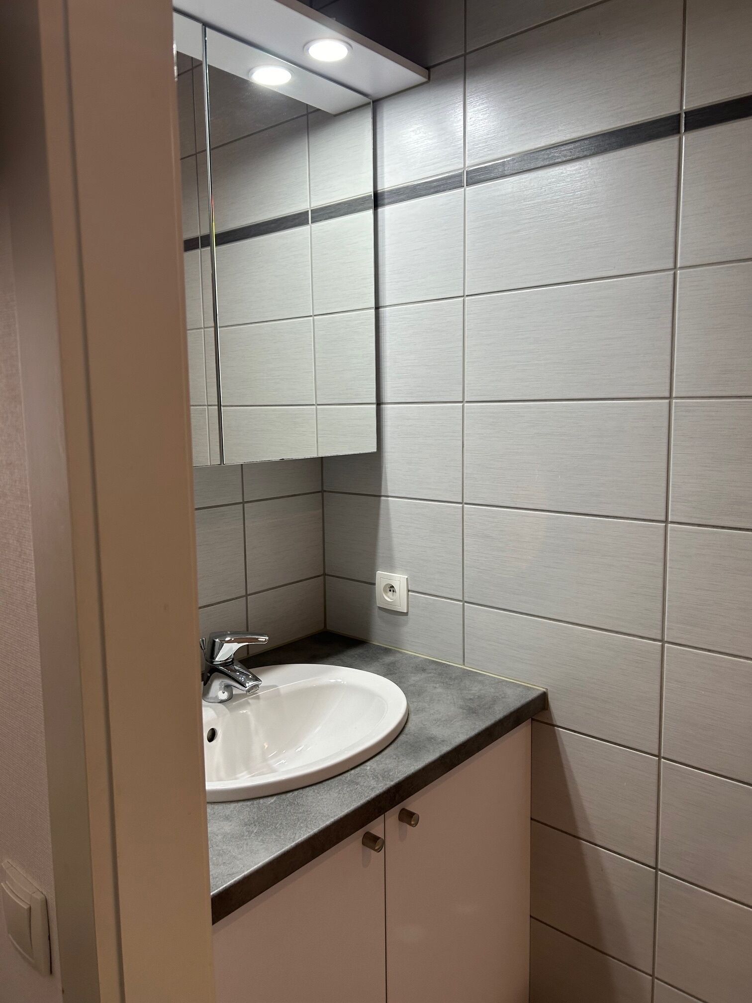Une salle de bain avec un lavabo blanc, un miroir et des murs recouverts de carreaux gris.