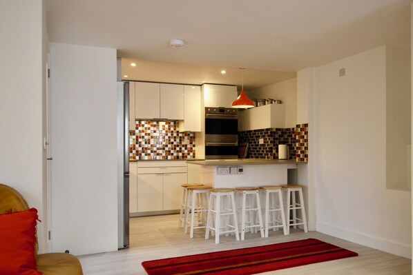 A modern kitchen with white cabinets, a red pendant light, and a tiled backsplash. There are four white stools at the counter. - Vernon Place Accommodation (Cheltenham)