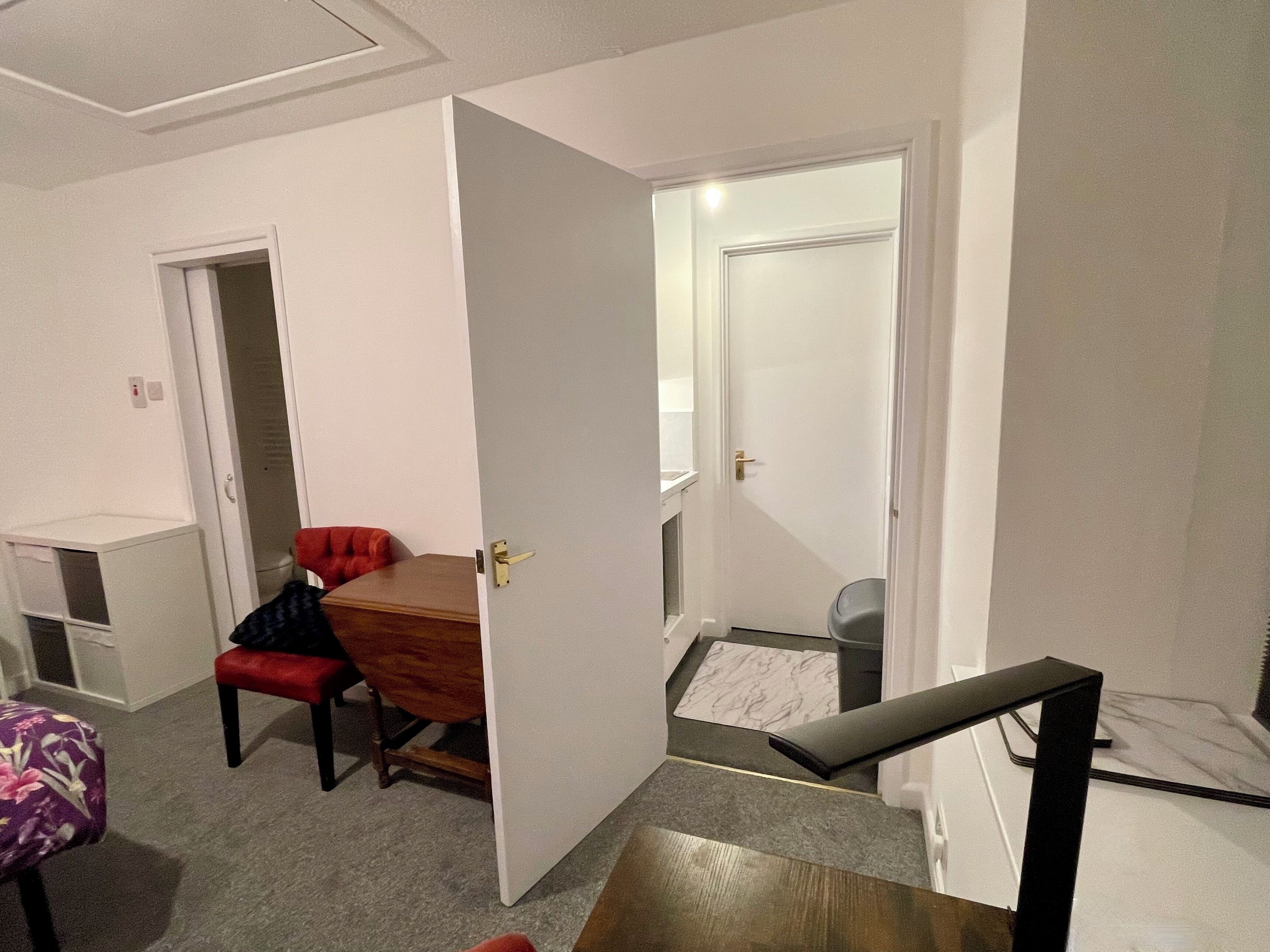 Une pièce de taille modeste, avec une table à manger en bois, une chaise rouge et un petit tabouret. Une porte donne accès à une salle de bain équipée de toilettes et d’une douche.