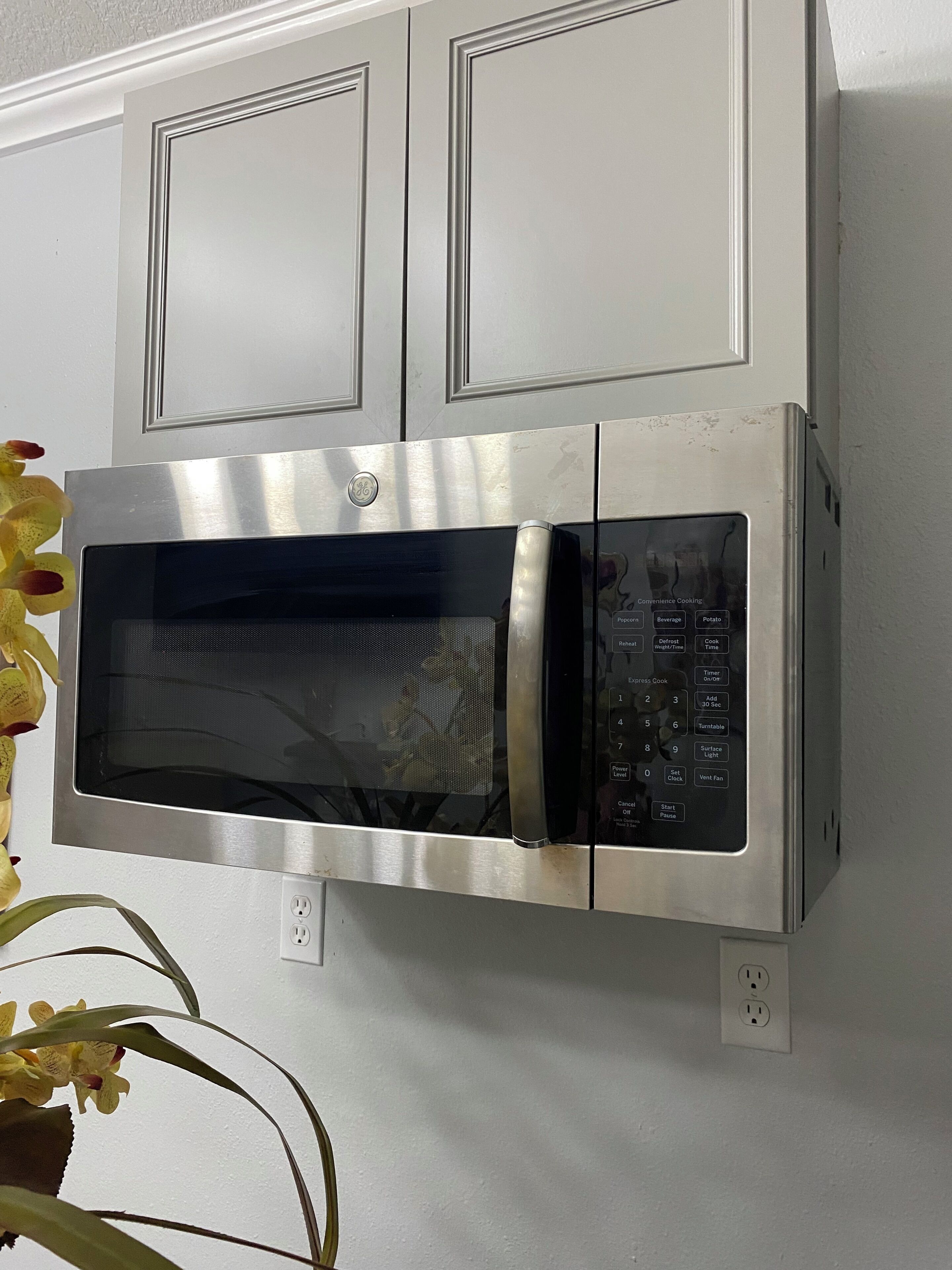 A wall-mounted microwave oven with a digital control panel, above a white wall with a power outlet.