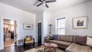 A spacious living room with a brown leather sofa, a coffee table, and a patterned rug. A dining area is visible in the background with a kitchen and a dining table.