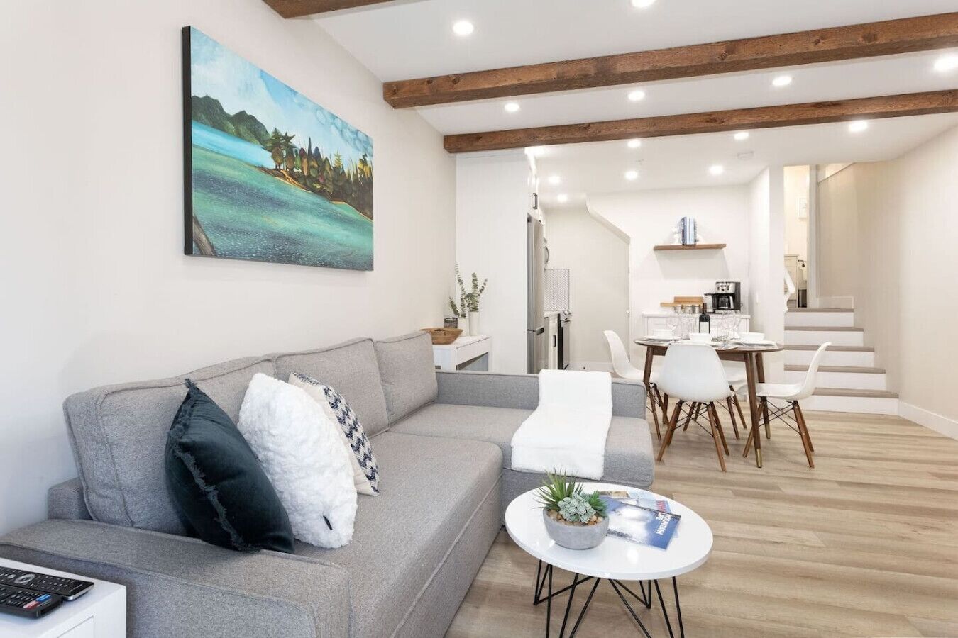 A modern living room with a grey sofa, white chairs, a small round table with a plant, and a painting on the wall.