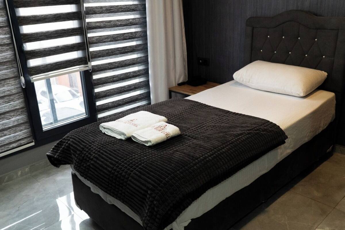 A hotel room with a bed, a nightstand, a window with blinds, and a view of the outside.