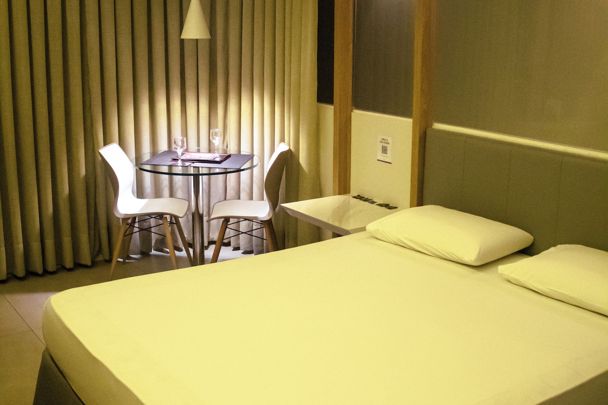 Habitación de hotel con una cama grande, una mesa redonda pequeña con dos sillas, y una lámpara colgante.