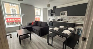A modern living room with a sofa, a dining table set for four, and a kitchen area with dark cabinets and a microwave.