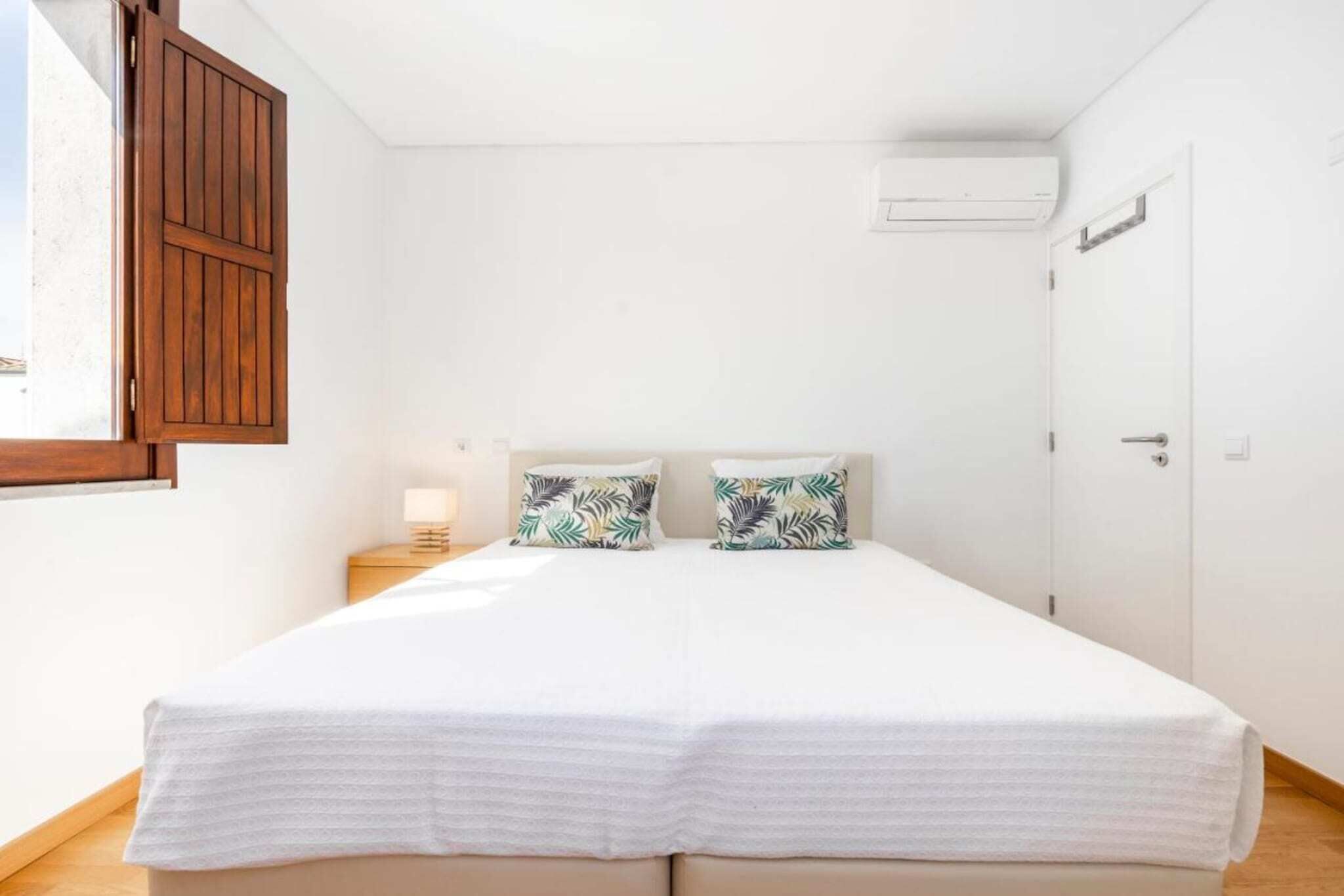 A bedroom with a double bed, two pillows with a tropical leaf pattern, a bedside table with a lamp, and a window with wooden shutters.