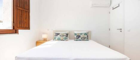 A bedroom with a double bed, two pillows with a tropical leaf pattern, a bedside table with a lamp, and a window with wooden shutters.
