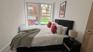 Um quarto com cama, almofadas, abajur, janela e porta.