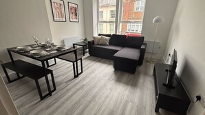 Uma sala de estar moderna com um sofá escuro, uma mesa de jantar para quatro pessoas e uma janela grande com vista para edifícios.