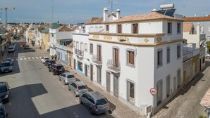 A white building with a tiled roof, multiple windows, and balconies. There are cars parked along the street in front. - Loft located in the city center with a large terrace and good sun exposure (Tavira)