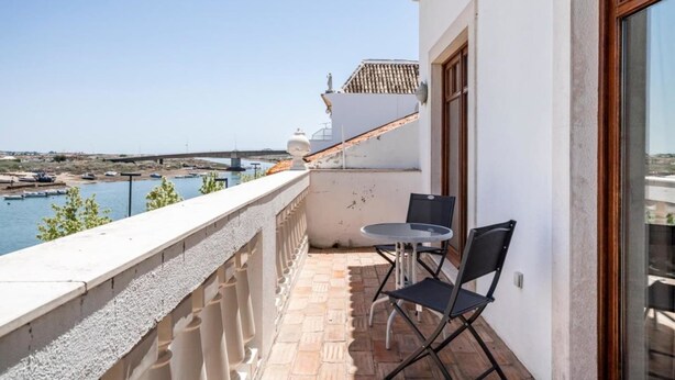 Un balcon avec une table et des chaises donnant sur un plan d’eau et un pont.