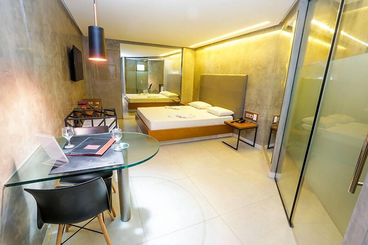 A modern hotel room with a glass dining table, a bed with a wooden headboard, and a small bedside table.