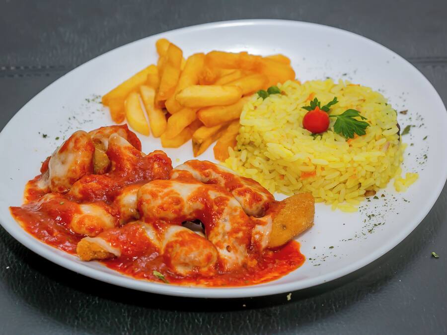 Un plato de pollo en salsa de tomate, papas fritas y arroz amarillo adornado con perejil y un tomate cherry.