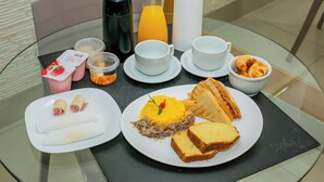 Ein Frühstückstisch mit verschiedenen Speisen, darunter gefüllte Teigtaschen, Reis mit Fleisch, Sandwiches und eine Schale Gebäck. Außerdem sind Tassen, ein Thermoskanne und ein Glas Orangensaft zu sehen.