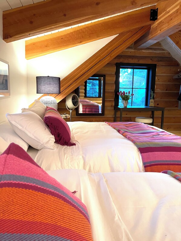 A cozy bedroom with a sloped ceiling, a bed with colorful bedding, a bedside lamp, and a window with a view of trees. - Luxury Log Cabin Retreat in Warm Springs, Sun Valley Idaho (Ketchum)
