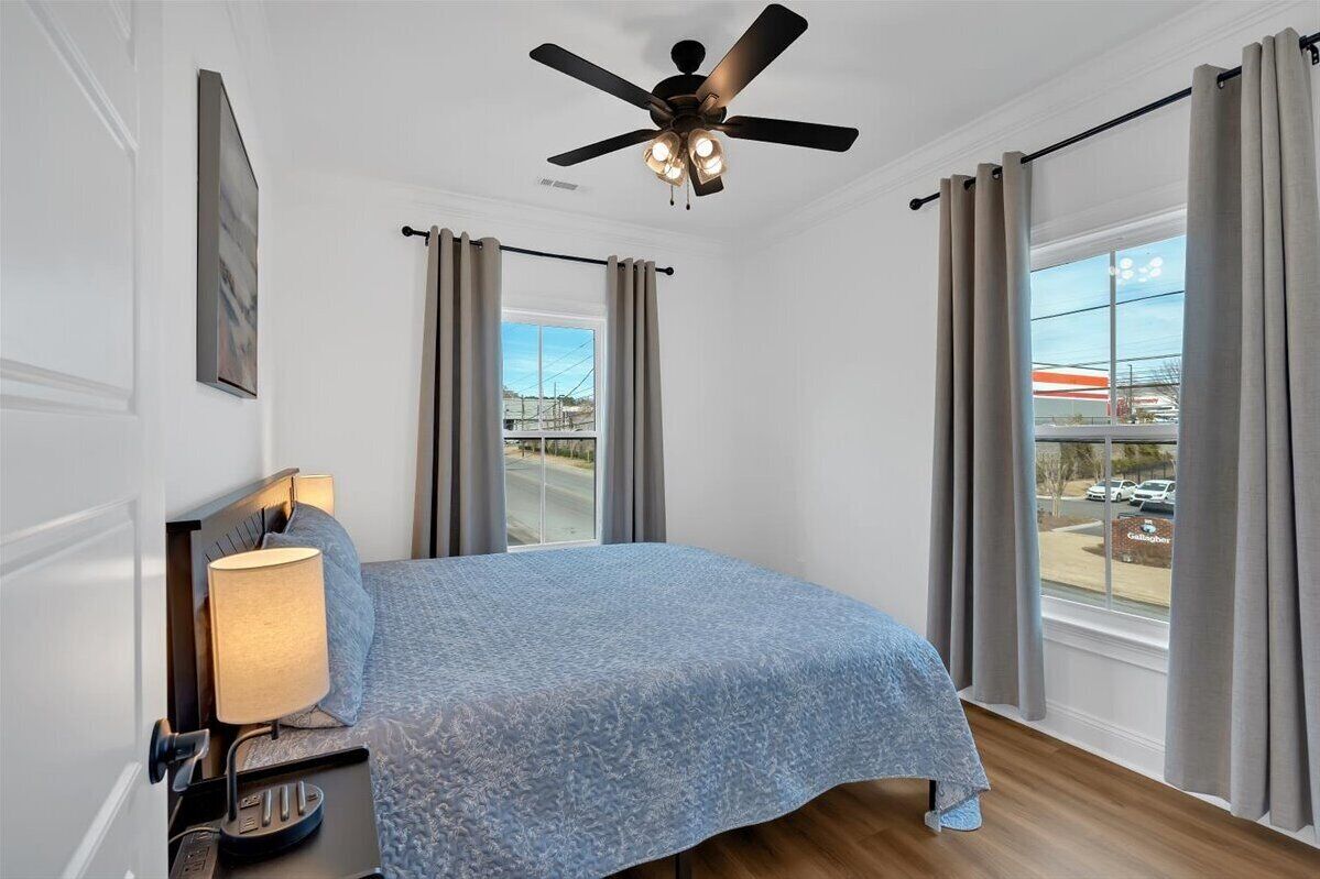 Une chambre d’hôtel avec un lit, un ventilateur de plafond, une table de chevet avec une lampe et de grandes fenêtres avec des rideaux.