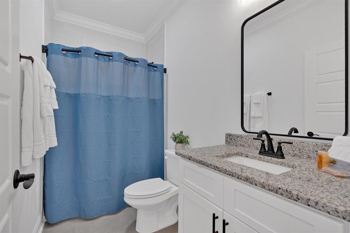 Une salle de bain avec des toilettes blanches, un rideau de douche bleu, un lavabo blanc avec un plan de travail en granit et un grand miroir.