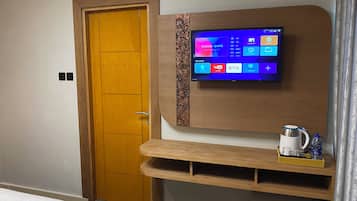 Una habitación de hotel moderna con una puerta de madera, un televisor montado que muestra varias aplicaciones, y una pequeña estantería con una tetera y una botella de agua.
