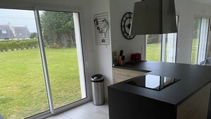 A modern kitchen with a large island, a stainless steel trash can, and a view of a green lawn through the sliding glass door. - Exceptional House for Rent in Finistère between Wild Beaches and Modern Comfort (Landunvez)