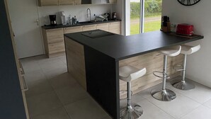 A modern kitchen with a central island, bar stools, and a large window providing a view of the outdoors. - Exceptional House for Rent in Finistère between Wild Beaches and Modern Comfort (Landunvez)