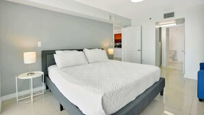 Habitación de hotel moderna con una cama grande, una mesita de noche con lámpara, y vistas al baño y la cocina.