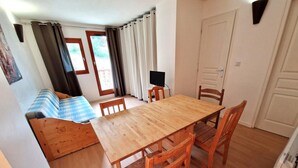 A cozy room with a wooden dining table, two wooden chairs, a small TV on a stand, a sofa with a checkered blanket, and a window with curtains. - 20m from ski slopes, Ground floor, view ski slopes, balcony, tv, ski locker, 32m², Valfréjus (Valfréjus)