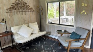 Une pièce chaleureuse avec un canapé blanc, un fauteuil gris, un tapis à motifs et une fenêtre donnant sur du mobilier d’extérieur.
