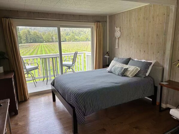 Un dormitorio con cama, una silla, un escritorio y vistas a un viñedo a través de una puerta corrediza.