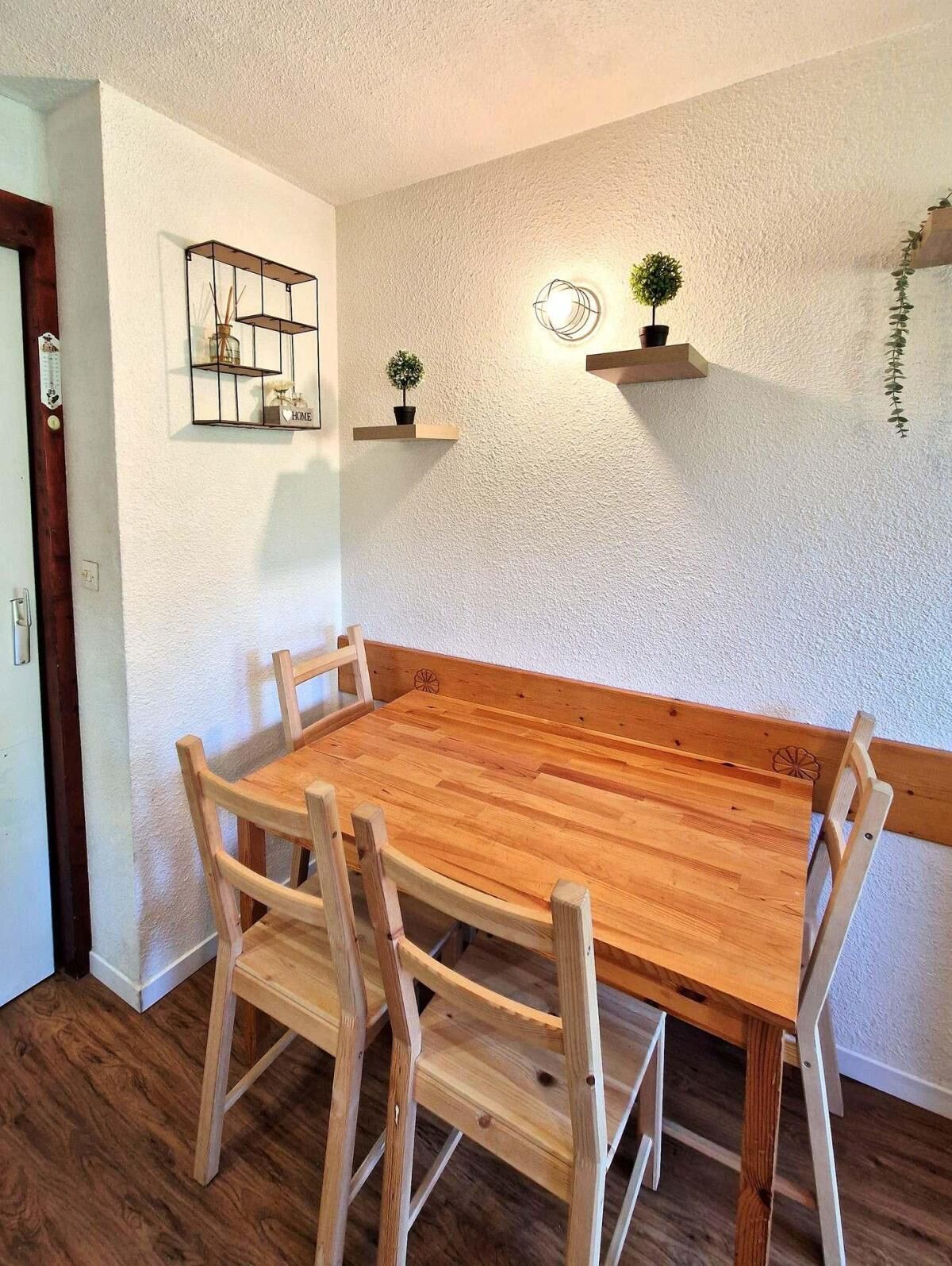 A dining area with a wooden table and chairs, a white textured wall, and two shelves with plants.