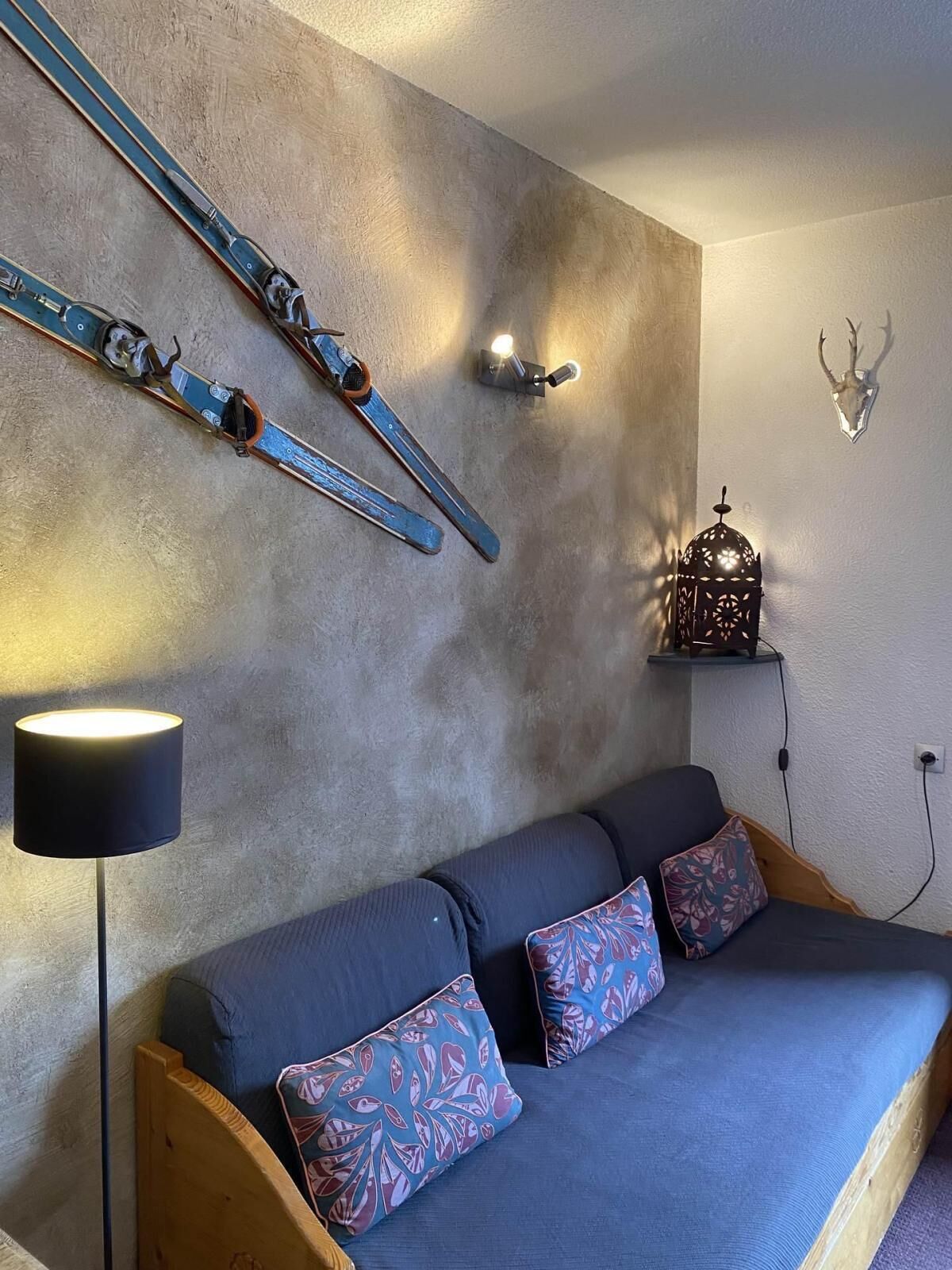 A cozy room with a grey sofa, patterned cushions, a wall-mounted ski rack, a wall-mounted light, and a decorative antler skull.