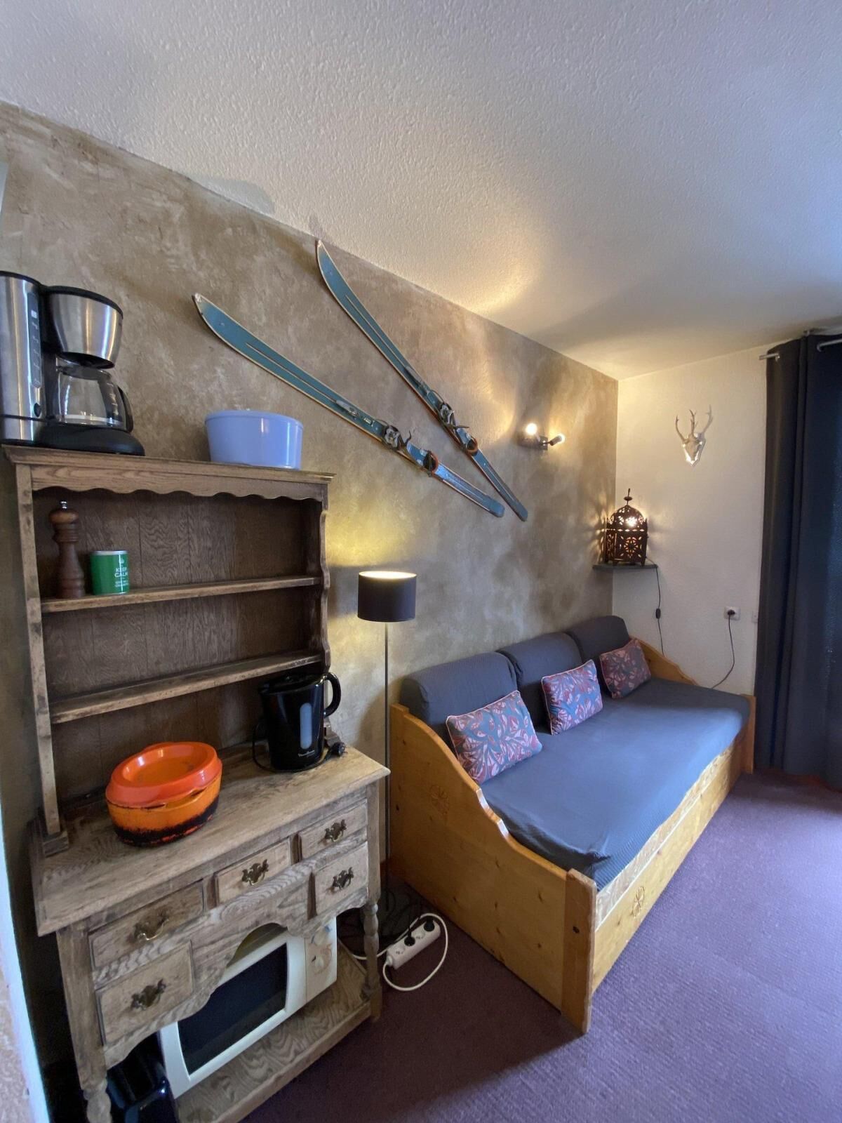 Een kleine kamer met een bed, een houten dressoir, een koffiezetapparaat, een kom en ski's aan de muur.