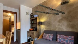 A cozy room with a wooden sofa, a dining table with chairs, and a kitchen area with a stove and shelves.