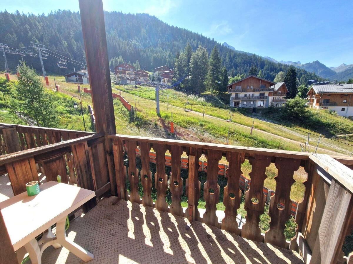 Ein Holzbalkon mit Tisch und Stühlen, der auf eine gebirgige Landschaft mit Häusern und einer Seilbahn blickt.