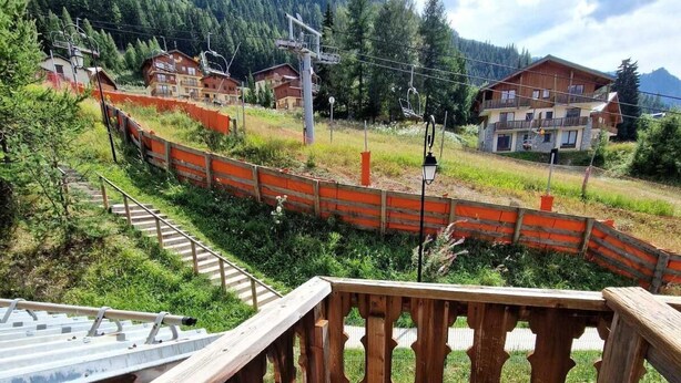A wooden balcony with a view of a mountainous landscape, a fenced area, and a building with multiple balconies.