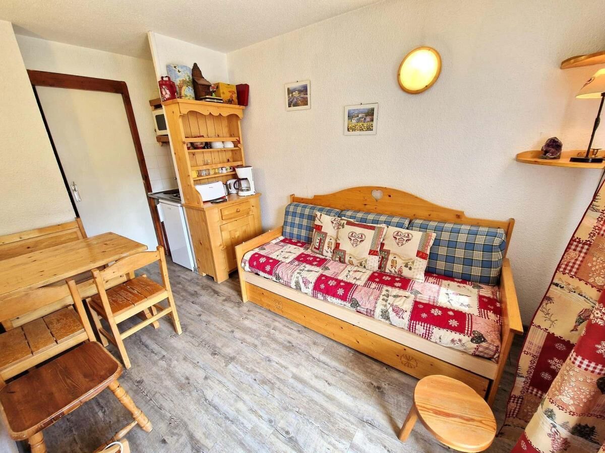 A cozy room with a wooden bed, a small wooden table, a chair, a shelf with kitchen items, and a wall-mounted lamp.