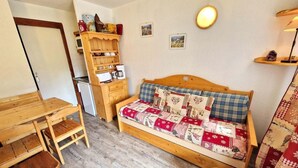 A cozy room with a wooden bed, a small wooden table, a chair, a shelf with kitchen items, and a wall-mounted lamp.